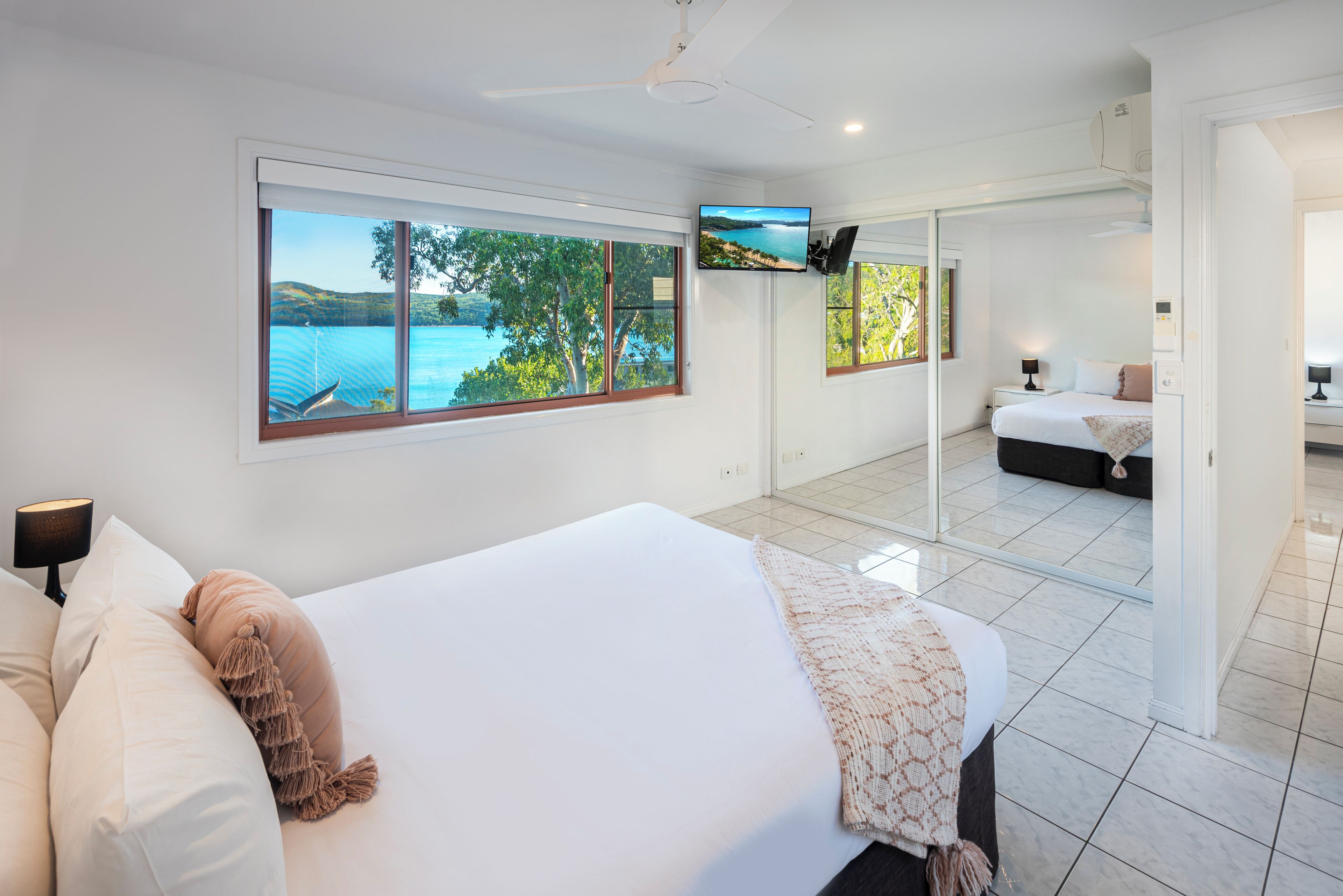 A bedroom with a large bed, a TV mounted on the wall, and a view of the sea through the windows.