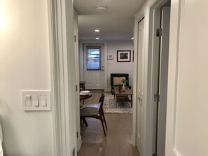 Un couloir mène à une salle à manger où se trouvent une table et des chaises, un canapé et une porte avec un interrupteur.