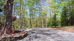 A paved driveway leading into a forested area with a large tree and a red wagon wheel. - A+ Location 2 mi Pigeon Forge Pkwy,Not up Mtn, Hottub, Quiet,Private,SUPER CLEAN (Sevierville)