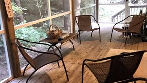 A wooden deck with wicker chairs and a table, large windows offering a forest view, and a 'Welcome' sign. - Hilltop Cottage in Pentwater Village (Pentwater)