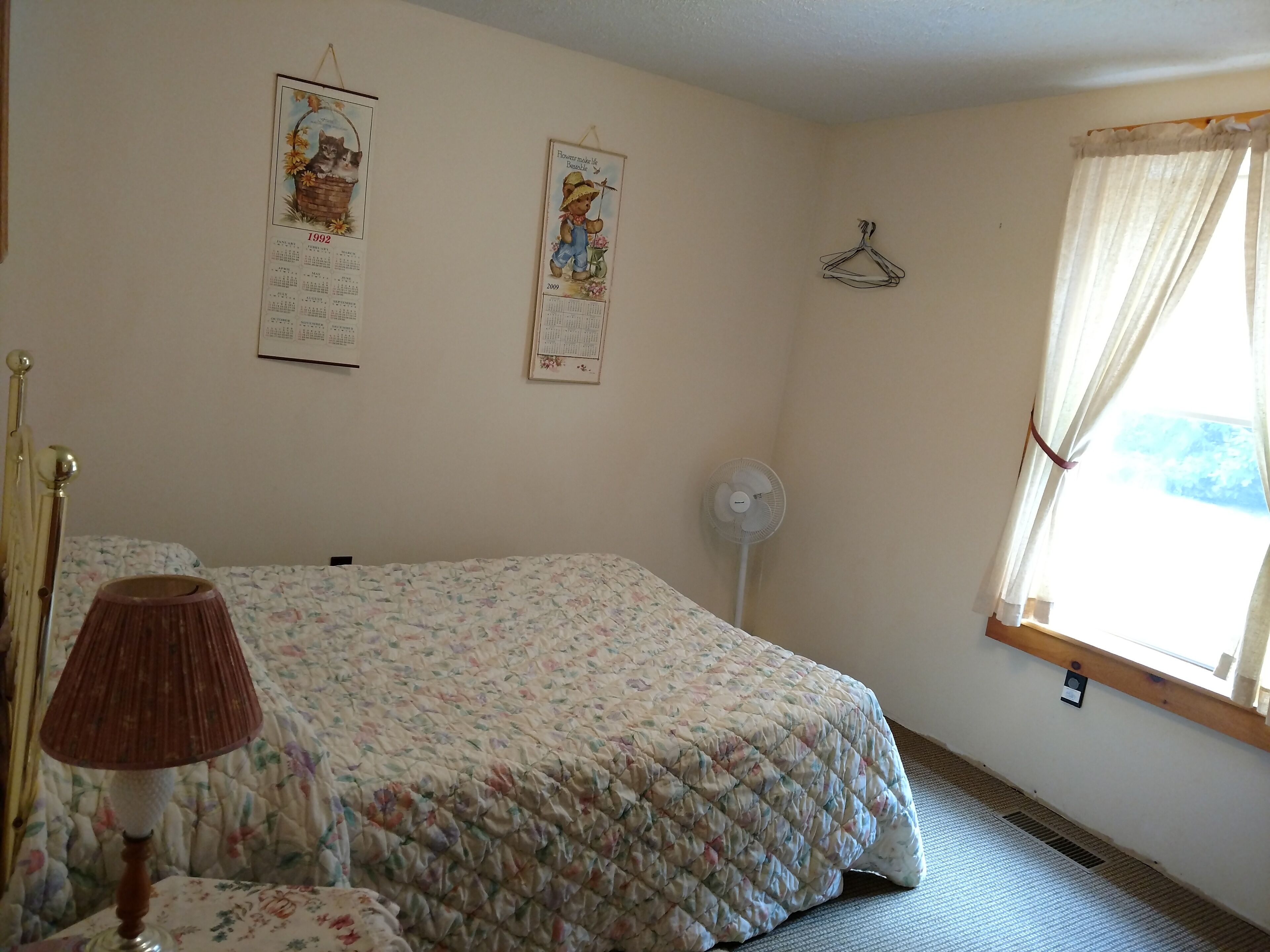 A bedroom with a floral bedspread, a bedside lamp, a wall calendar, a hanging coat rack, and a window with curtains.