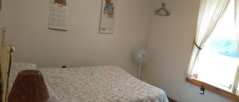 A bedroom with a floral bedspread, a bedside lamp, a wall calendar, a hanging coat rack, and a window with curtains.