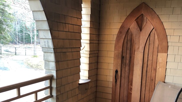 Una esquina de una habitación con pared de ladrillo, una puerta de madera y una ventana. Hay una silla y una mesa con una lámpara.