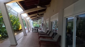 A covered outdoor seating area with tables and chairs, a ceiling fan, and large windows.