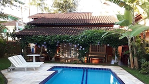A rectangular swimming pool with blue tiles, surrounded by a tiled patio. There is a white lounge chair and a table with chairs outside the pool area.