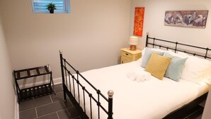 Ein Schlafzimmer mit schwarzem Metallbettgestell, weißer Bettdecke und mehreren Kissen. Ein kleiner Nachttisch mit Lampe, ein Fenster mit Pflanze und ein Gemälde an der Wand sind ebenfalls zu sehen.