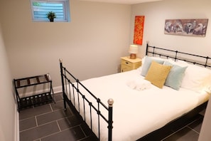 A bedroom with a black metal bed frame, a white bedspread, and multiple pillows. There is a small bedside table with a lamp, a window with a plant, and a painting on the wall.