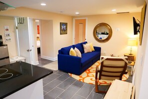 A compact living space with a kitchenette, a blue sofa, a wooden chair, and a round mirror on the wall.