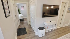 Un couloir avec une porte blanche, un téléviseur blanc fixé au mur, un bureau blanc avec une télécommande et un radiateur blanc.