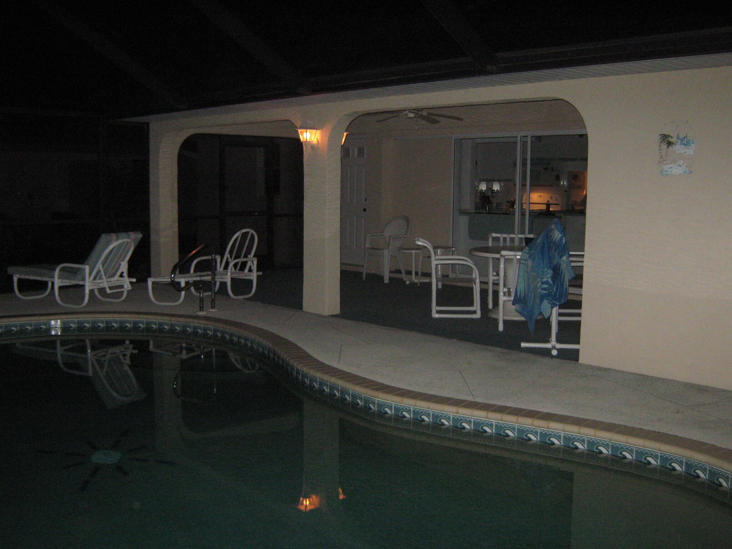 A pool area with chairs and a table, a ceiling fan, and a kitchen visible through an open doorway.