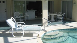 Uma área de piscina com espreguiçadeiras e uma mesa, um alpendre coberto com cadeiras, e uma porta que dá para um espaço interno.
