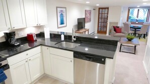 Una cocina moderna con armarios blancos, lavavajillas de acero inoxidable, encimera negra y un comedor con mesa y sillas.