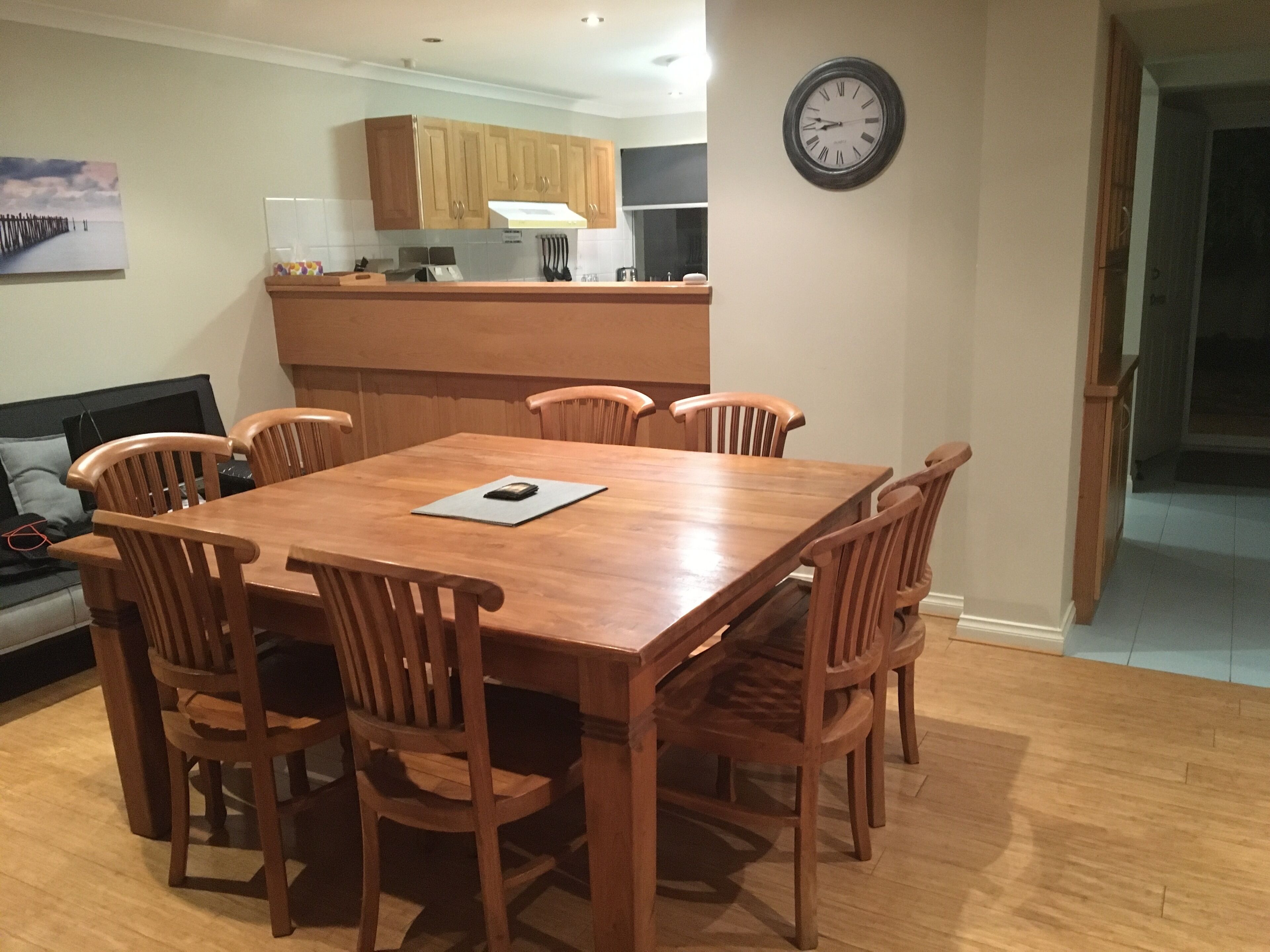 Un coin repas avec une table et des chaises en bois, une cuisine à l’arrière-plan et une horloge au mur.