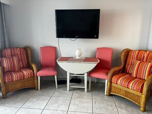 A room with two wicker chairs with red cushions, a small white table with a vase, and a flat-screen TV mounted on the wall.