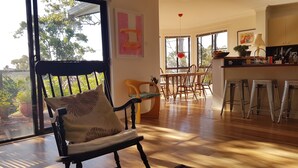 Un vaste espace de vie doté d’un plancher en bois, d’une table à manger avec des chaises, d’un coin cuisine avec des tabourets et d’une grande fenêtre offrant une vue sur la verdure.