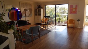 Un salon spacieux doté d’un plancher en bois, d’une table à manger, d’une chaise bleue, d’un fauteuil à bascule et d’une grande porte-fenêtre coulissante donnant sur un balcon agrémenté de plantes en pot.
