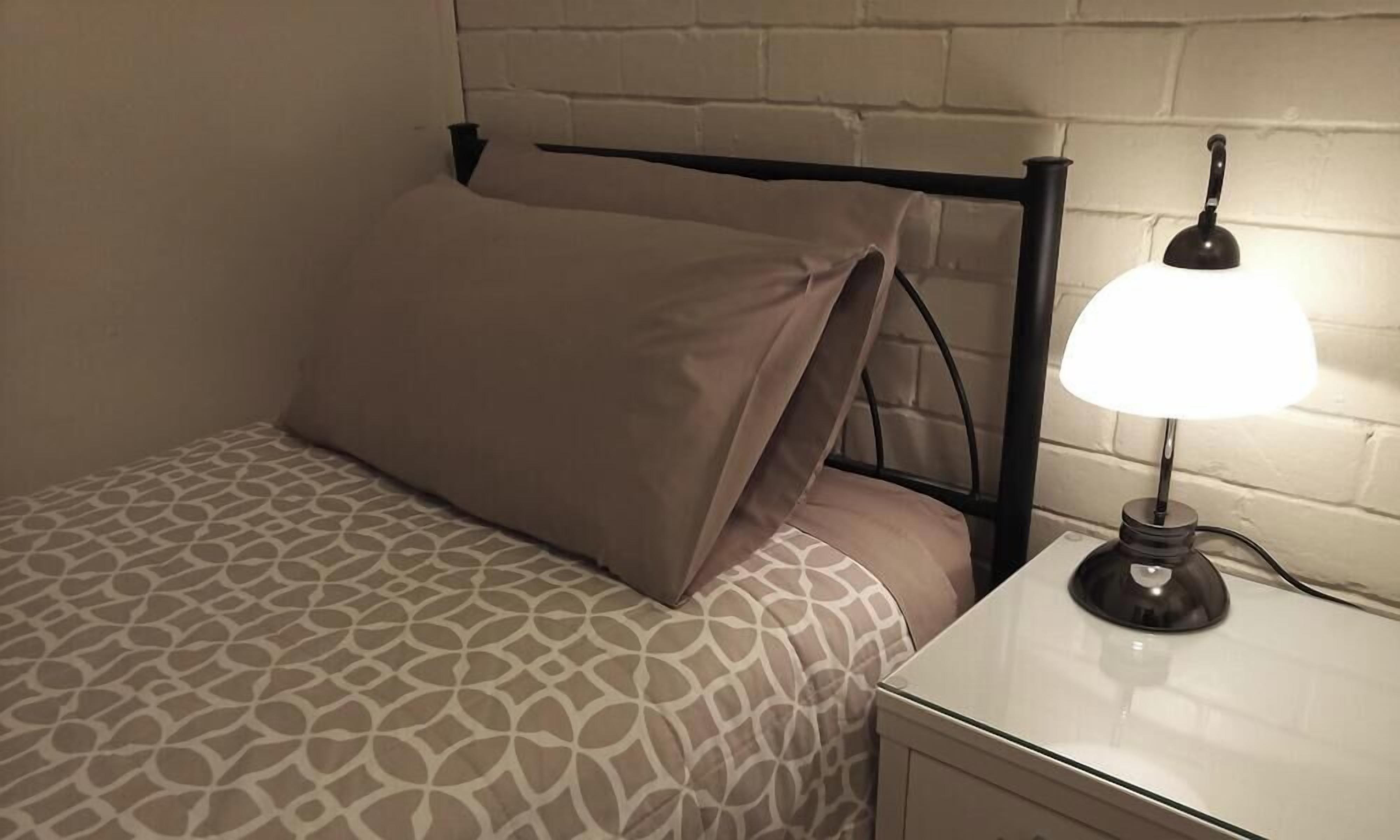 A bed with a patterned bedspread, a single pillow, and a bedside lamp on a white nightstand.