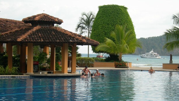 A pool area with a gazebo, palm trees, and a view of the sea. - Los Suenos Marina and Resort - Del Mar Luxury Condo 3 Br/2 Ba (Herradurra Beach Jaco)