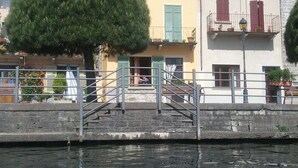A waterfront building with a staircase leading to a balcony, a tree in front, and a calm water body reflecting the buildings. - Pella: Pella, Lake of Orta: house on the Lake,fantastic view (Pella)