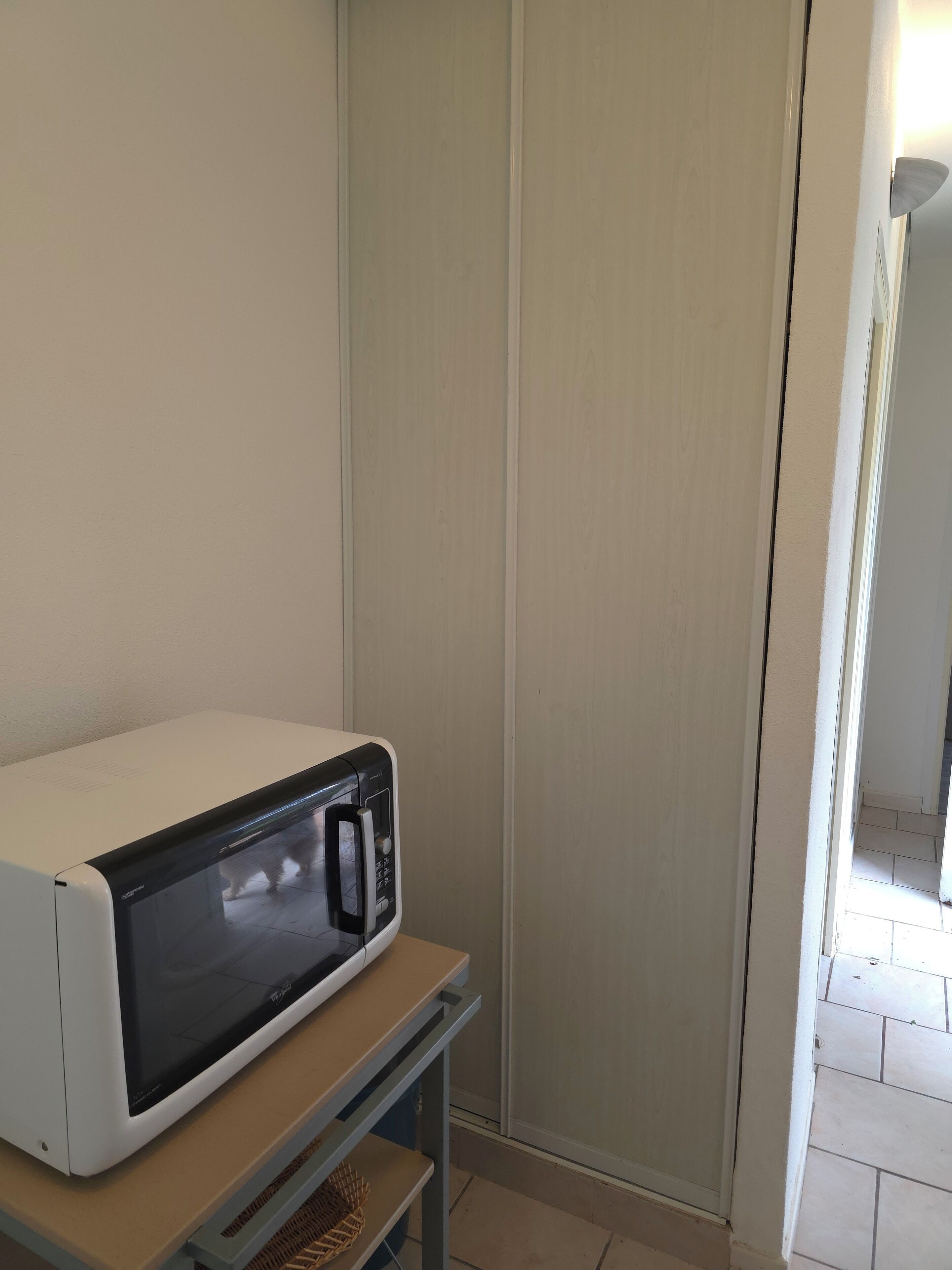Un four à micro-ondes blanc est posé sur une table en bois dans une pièce aux murs de couleur claire, et une porte donne accès à une autre pièce.