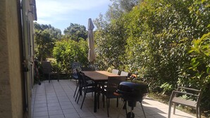 An outdoor patio with a tiled floor, a wooden table, black chairs, a barbecue grill, and a covered umbrella.
