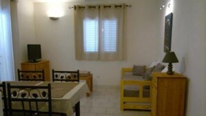 A small, well-lit room with a dining table and chairs, a sofa, a TV on a wooden cabinet, and a window with curtains.