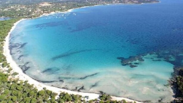 An aerial view of a coastal area with a clear blue sea, sandy beach, and surrounding greenery.