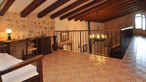 Une chambre spacieuse aménagée dans un grenier, avec des poutres apparentes, un mur en pierre, une grande fenêtre et un escalier doté d’une rampe en fer forgé.