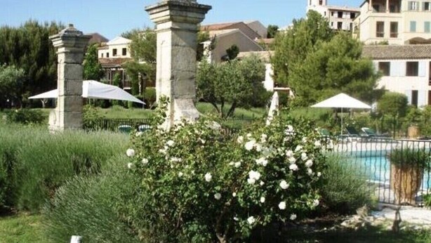 A pool area with a fountain, surrounded by greenery and a building in the background. - Mas de Pont Royal - Golf Estate - Pool & Garden (Mallemort)