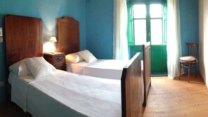 A bedroom with two beds, a wooden wardrobe, a nightstand with a lamp, a chair, and a window with curtains. - The Ancient House, wonderful Sardinia! (Masainas)