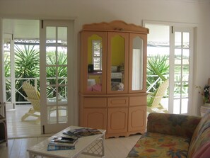 A room with a wooden cabinet, a sofa, a small table with magazines, and a view of a balcony with chairs and plants. - Gingerbread Cottage 2 bed with pool, seaviews, Nr Holetown St. James (Mount Standfast)