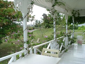 A covered porch with white railings and a bench, surrounded by greenery and flowers. - Gingerbread Cottage 2 bed with pool, seaviews, Nr Holetown St. James (Mount Standfast)