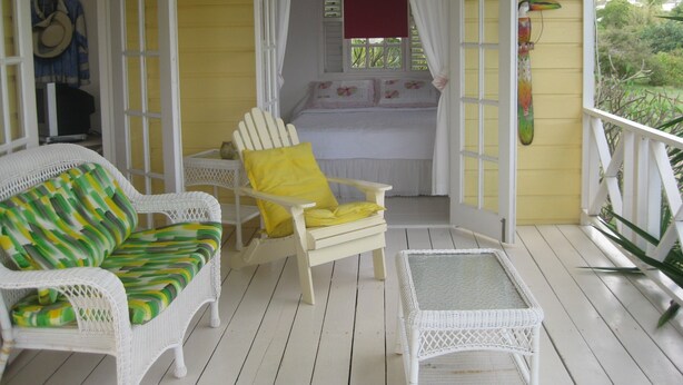 A room with a bed, a white wicker chair with a yellow pillow, a wicker sofa with a green and yellow striped cushion, and a small white table.