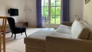 Un salon avec un canapé beige, une chaise en bois, un téléviseur, une lampe et une fenêtre avec des rideaux.