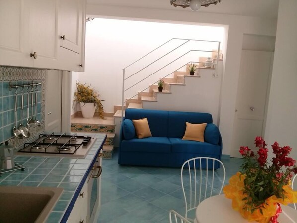 A modern kitchen with a blue tiled backsplash, a gas stove, and white cabinets. There is a blue sofa and a white chair in the living area, with a staircase leading to an upper floor.