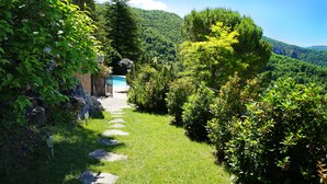 A stone pathway leading to a pool surrounded by lush greenery and a mountainous backdrop. - Stunning charming detached house with private heated pool (La Roque-Sainte-Marguerite)