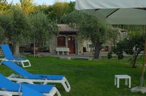 A garden with blue lounge chairs, a stone building with a wooden door, and a white umbrella. - Bosa: House / Villa - ModoloBig house (Bosa)