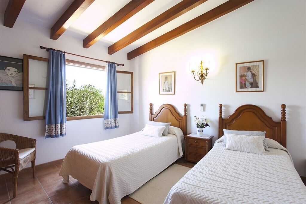 Une chambre avec deux lits simples, des têtes de lit en bois, une table de chevet, une chaise en osier et une fenêtre avec des rideaux bleus.
