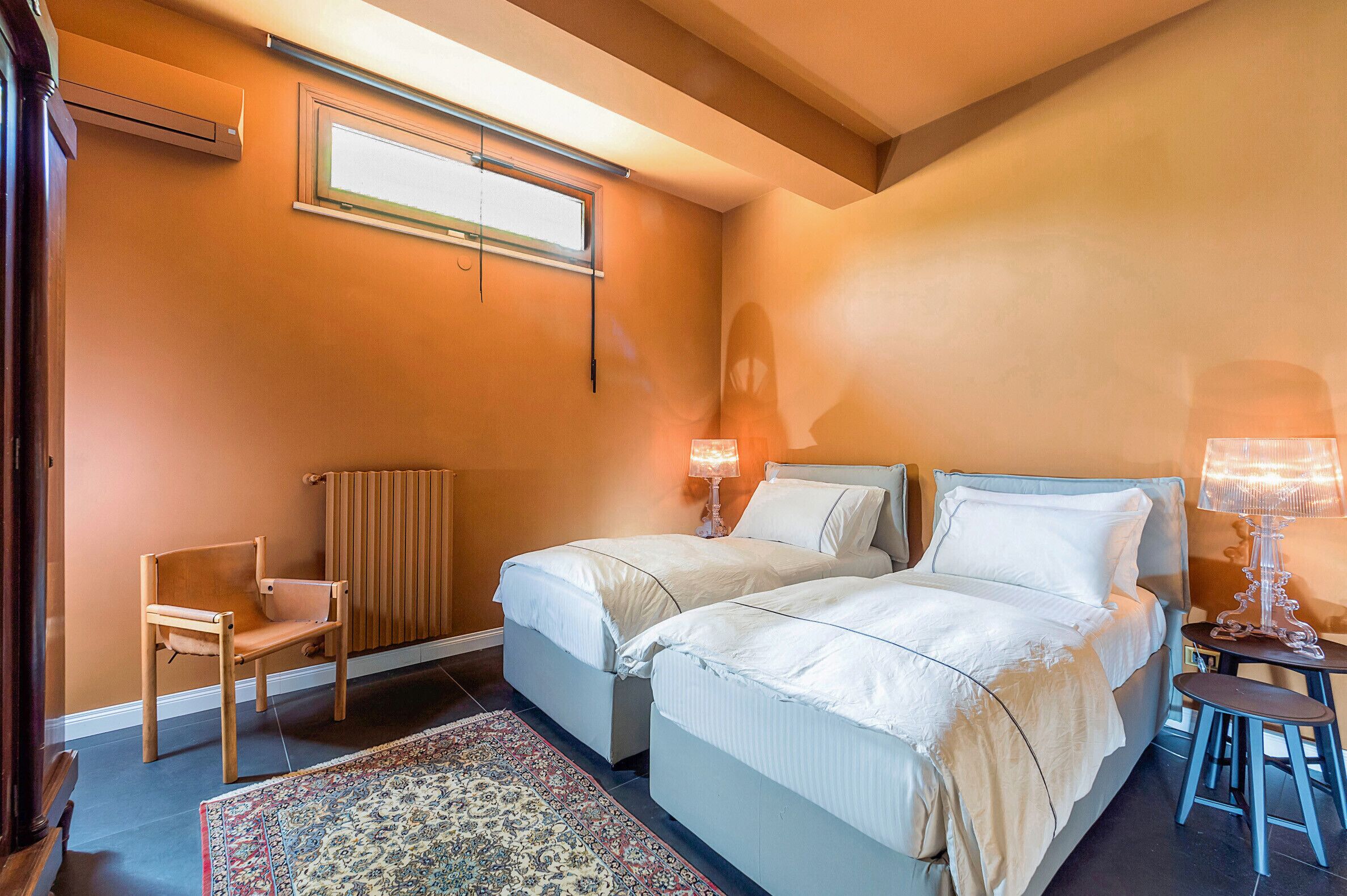 A bedroom with two beds, a chair, a radiator, and a small table with a lamp.