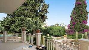 Un balcon donnant sur une piscine, un cactus et un grand arbre aux fleurs violettes.