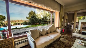 A room with a large window overlooking a pool and garden, a sofa, and a dining area with a table and chairs.