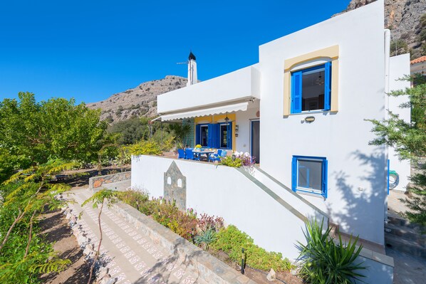 Un edificio blanco con detalles azules, un camino de piedra y montañas al fondo.