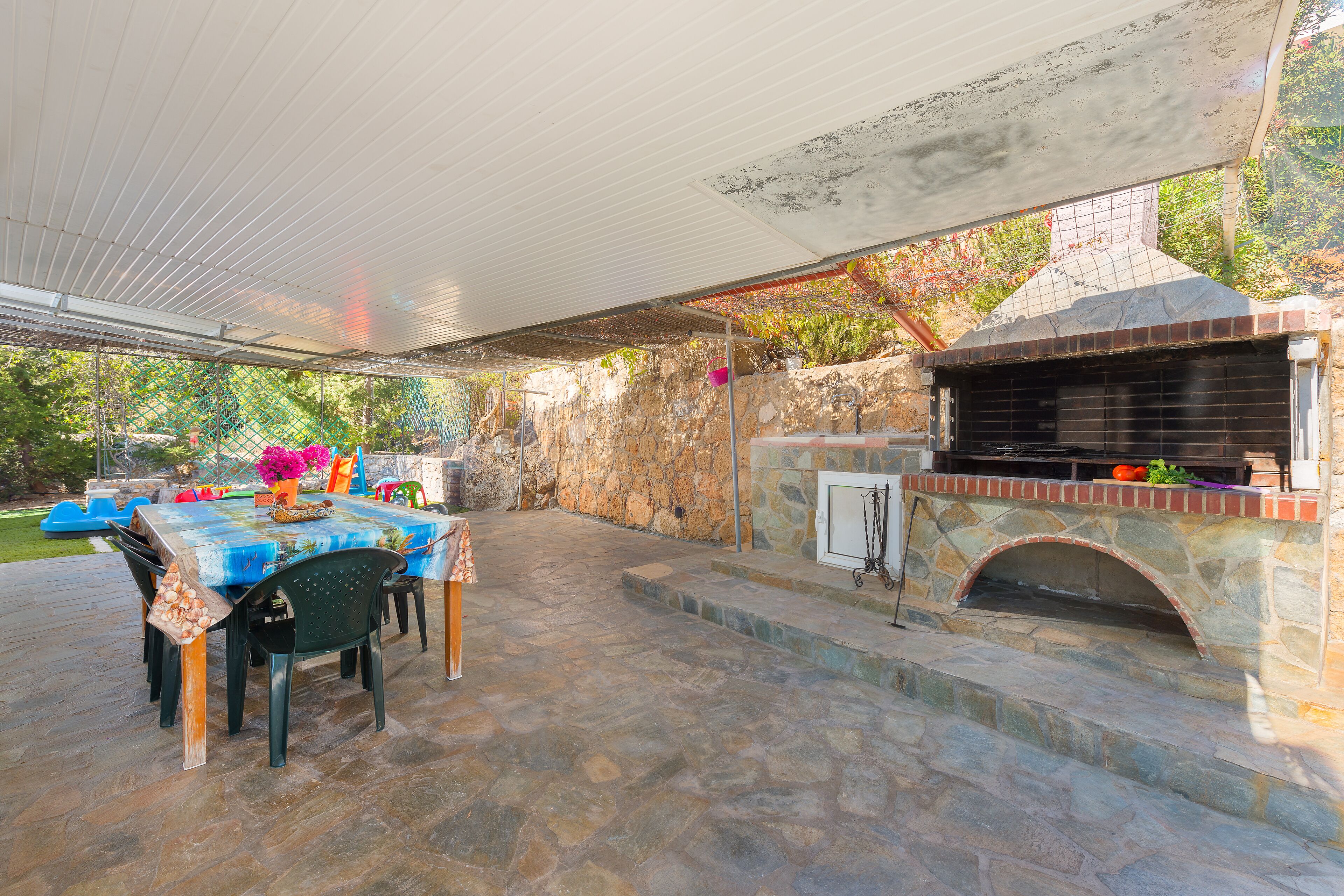 An outdoor patio with a stone table and chairs, a brick oven, and a covered area.