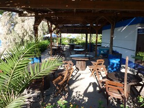 Un patio al aire libre con mesas y sillas de madera, rodeado de plantas en macetas y una zona cubierta para sentarse.
