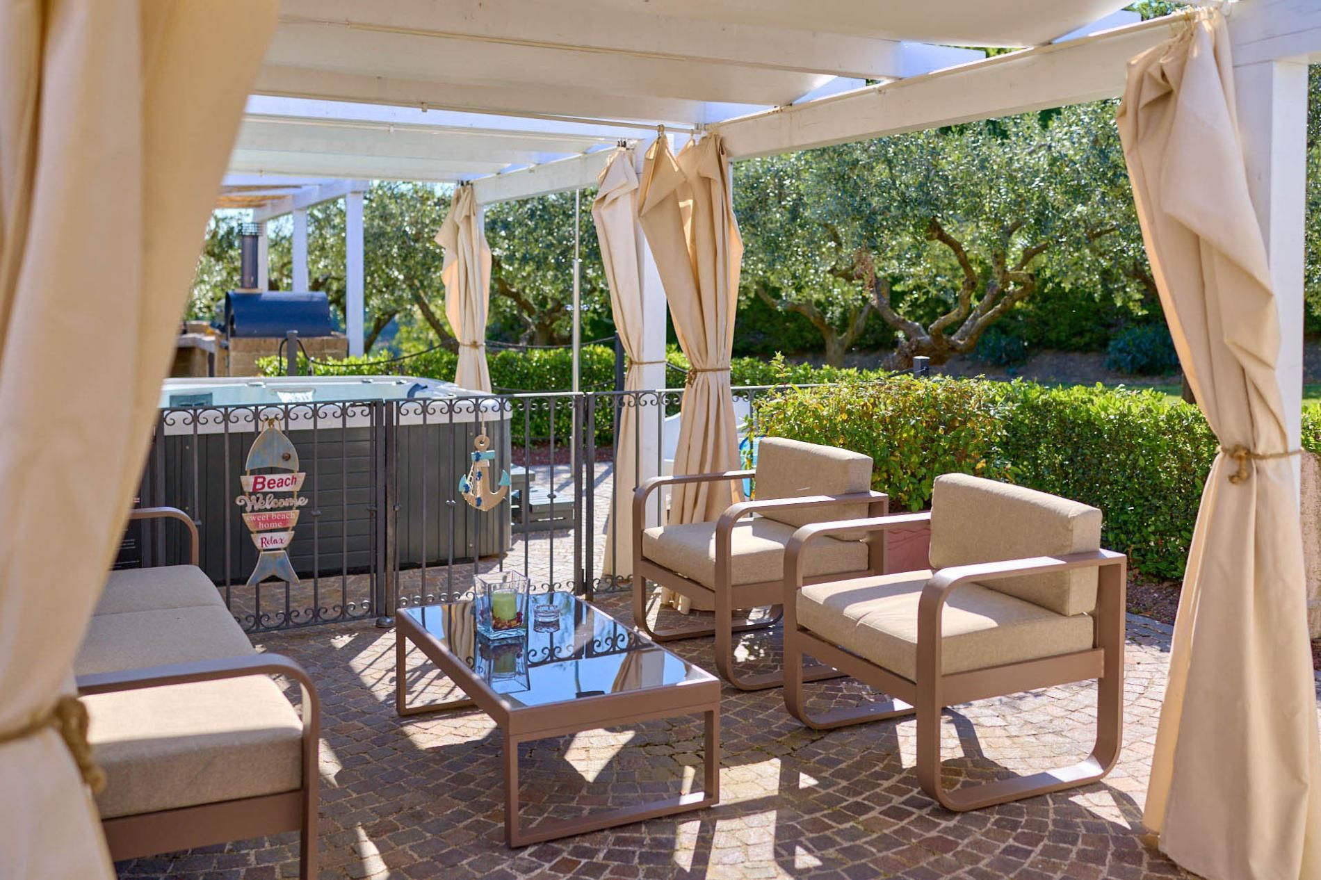 Ein Außenbereich mit einer Terrasse, einem Tisch mit Glasplatte, beigefarbenen Sitzgelegenheiten und einem Grill.