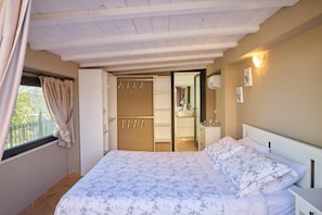 Ein Schlafzimmer mit Bett, Kleiderschrank, einem durchgehenden Türspalt sichtbaren Badezimmer und einem Fenster mit Vorhängen.