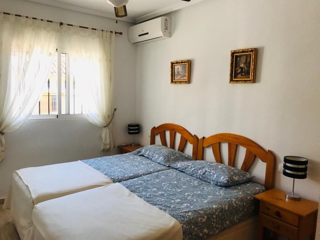 A bedroom with two beds, a window with sheer curtains, two framed pictures on the wall, and a bedside table with a lamp.
