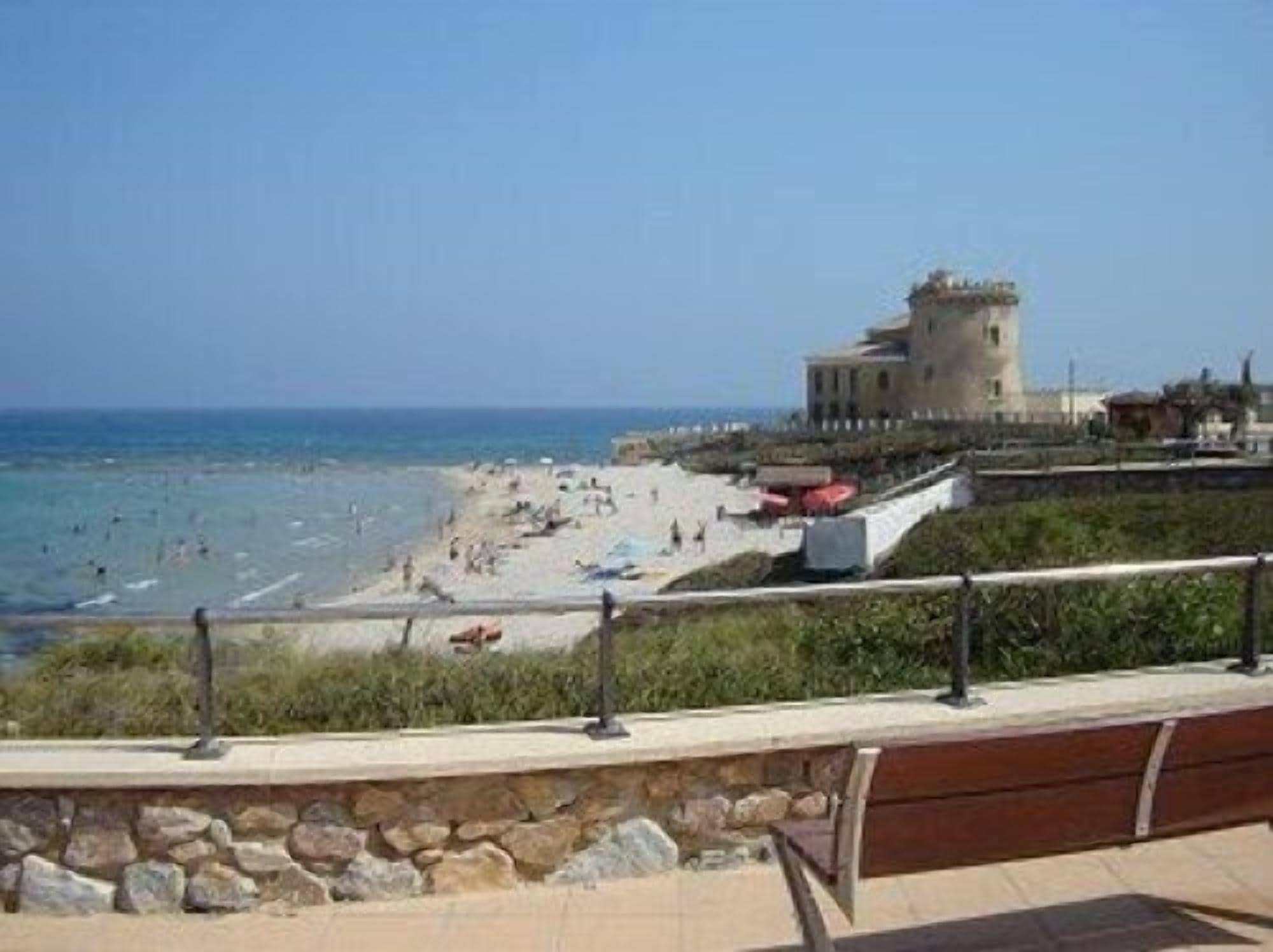 A coastal view with a castle-like structure, beach, and a stone wall with a bench.