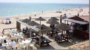 Ein Restaurant direkt am Strand mit Außenbereich, inklusive Tischen, Stühlen und reetgedeckten Sonnenschirmen.