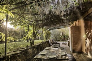 An outdoor dining area with a stone wall, wooden table, and chairs, surrounded by greenery and a pergola.